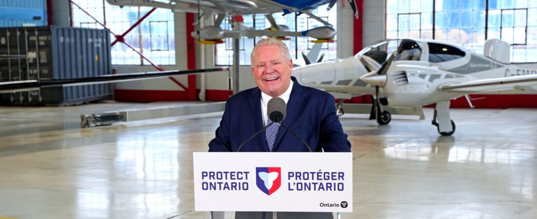 Doug Ford fala ao microfone num hangar, com aviões ao fundo.