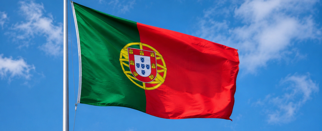 Bandeira de Portugal hasteada ao vento sob um céu azul com algumas nuvens.